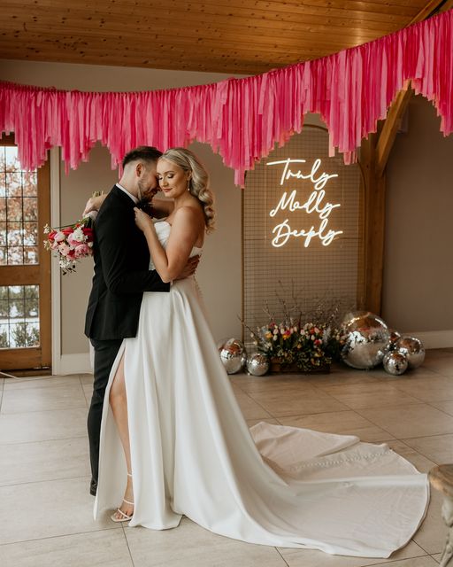 This is a photo of a female bride and a male groom embracing with a custom LED Neon in the background that they have hired from The Word is Love in England